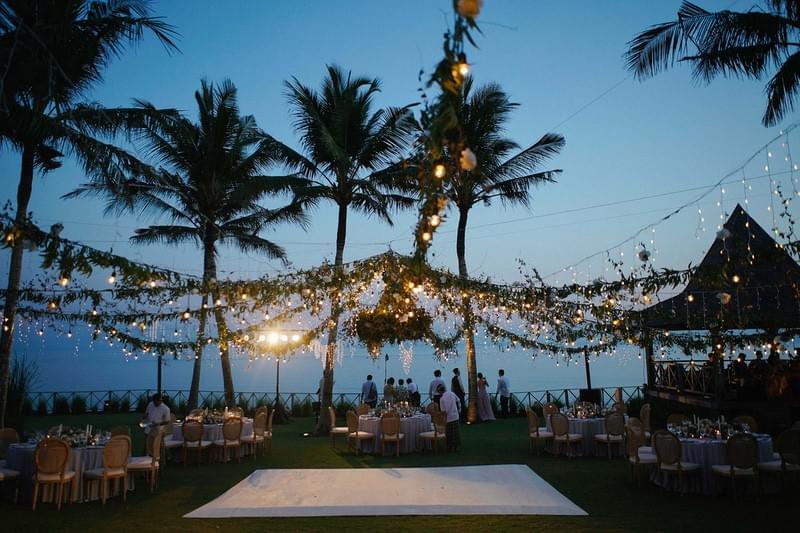  Magical outdoor wedding reception at Khayangan Estate during twilight, featuring a white dance floor, round dining tables with elegant seating, string lights, lush greenery, and an ocean backdrop framed by palm trees 
