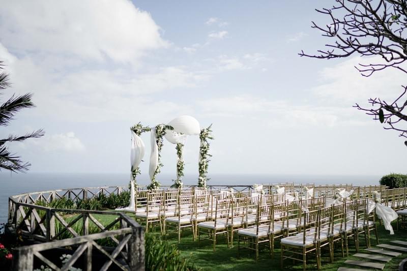  Clifftop wedding ceremony setup at Khayangan Estate, featuring a square floral arch with draped white fabric, gold Chiavari chairs, and a stunning ocean backdrop 