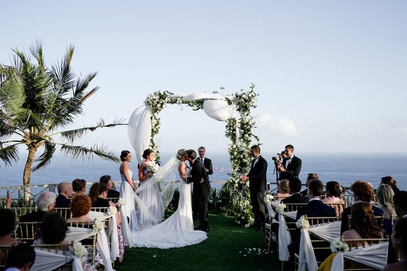  Romantic clifftop wedding ceremony at Khayangan Estate, featuring a draped floral arch, gold chairs with white accents, and elegantly dressed guests enjoying panoramic ocean views 