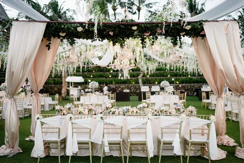  Elegant bridal table setup at Khayangan Estate, featuring gold chairs with draped white sashes, pastel floral arrangements, and a canopy adorned with cascading greenery, flowers, and chandeliers 