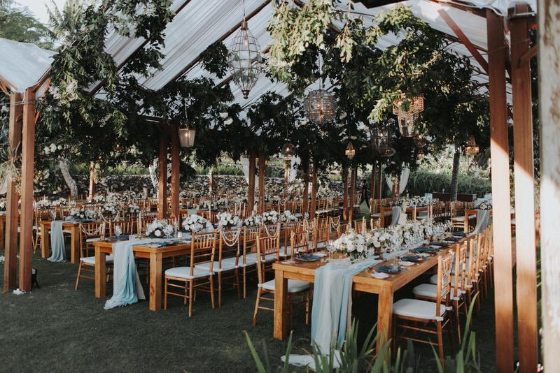  Rustic wedding reception setup at Khayangan Estate, featuring long wooden tables with flowing table runners, gold Chiavari chairs, floral centerpieces, and lantern lighting under a canopy of greenery 