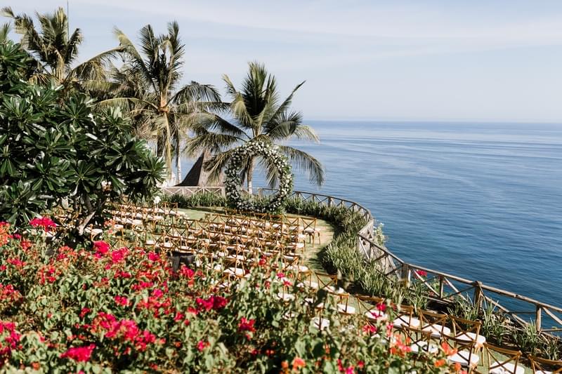  Clifftop wedding ceremony setup at Khayangan Estate, featuring a circular floral arch, wooden cross-back chairs, and vibrant bougainvillea overlooking the ocean. 