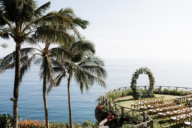  Aerial view of a clifftop wedding ceremony setup at Khayangan Estate, featuring a circular floral arch, wooden cross-back chairs, and lush palm trees overlooking the ocean 