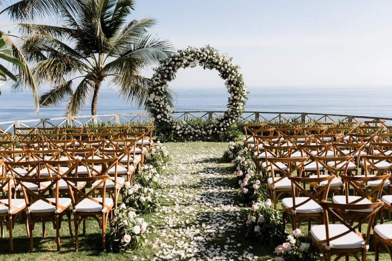 Wedding ceremony setup at Khayangan Estate, featuring wooden cross-back chairs, a circular floral arch, and a petal-lined aisle on a clifftop lawn overlooking the ocean 