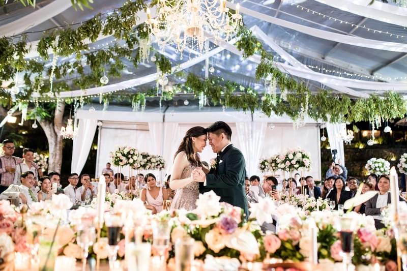  Bride and groom share a romantic first dance under a clear marquee at Khayangan Estate, surrounded by elegantly decorated tables, floral arrangements, chandeliers, and their seated guests 