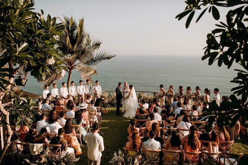  Intimate wedding ceremony at Khayangan Estate, set on a clifftop lawn with panoramic ocean views, natural floral arrangements, and wooden cross-back chairs for guests. 