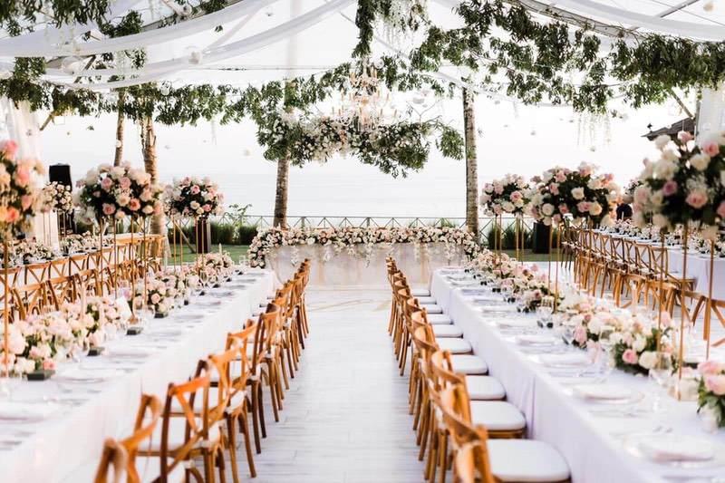  Luxurious wedding reception at Khayangan Estate, featuring long banquet tables adorned with pink and white floral arrangements, wooden chairs, hanging greenery, and chandeliers under a clear marquee with an ocean backdrop 