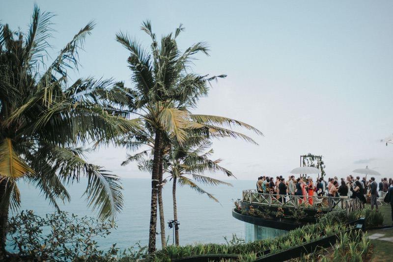  Aerial view of a clifftop wedding ceremony at Khayangan Estate, featuring a floral arch, elegantly dressed guests, and lush palm trees overlooking the ocean 