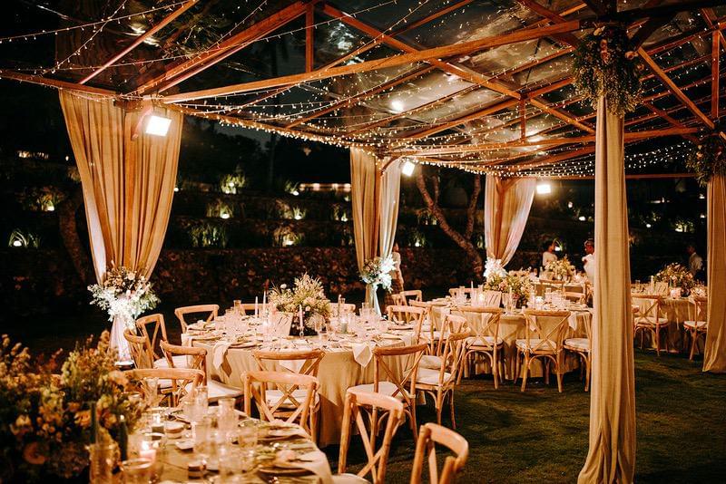  Elegant outdoor wedding reception at Khayangan Estate, featuring round tables with cross-back wooden chairs, soft draped columns, fairy lights, and a transparent canopy under the night sky 