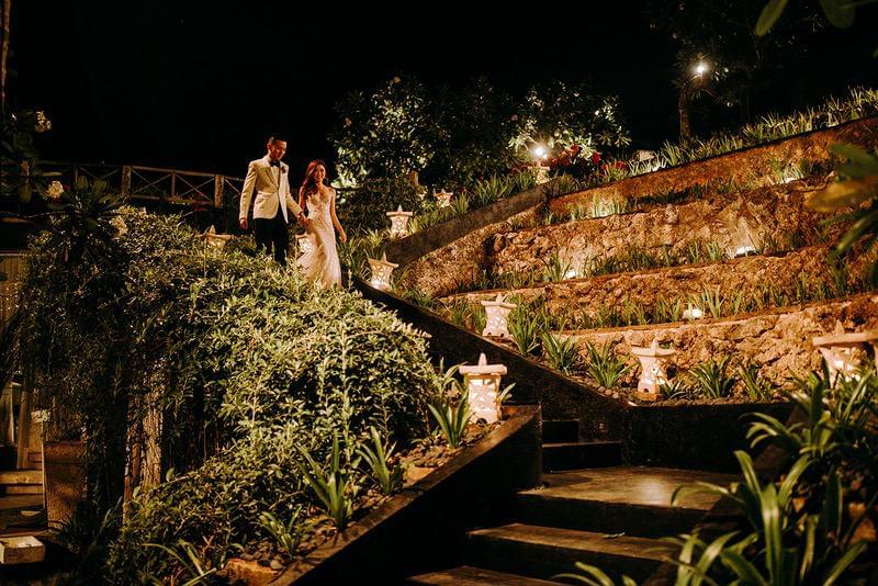  Bride and groom walking hand in hand down beautifully lit stone steps surrounded by lush greenery during their evening wedding at Khayangan Estate 