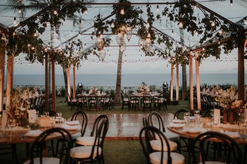  Outdoor wedding reception at Khayangan Estate under a clear marquee, featuring a central wooden dance floor surrounded by wooden round tables, black bentwood chairs, string lights, hanging lanterns, and an ocean backdrop 