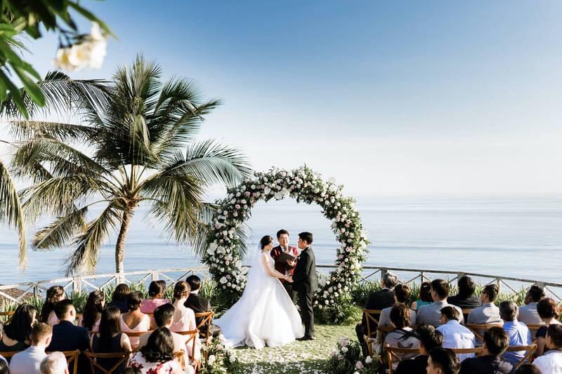  Bride and groom exchange vows under a circular floral arch during a clifftop wedding ceremony at Khayangan Estate, surrounded by seated guests and overlooking the ocean. 