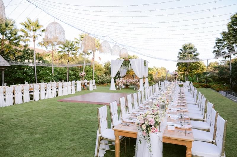  Elegant outdoor wedding reception setup at the Senses Lawn of The Ritz-Carlton Bali, featuring long banquet tables adorned with pastel floral arrangements and string lights overhead 