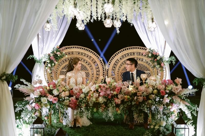 Elegant bridal table adorned with lush floral arrangements and romantic lighting, featuring the bride and groom seated under a beautifully draped canopy. 