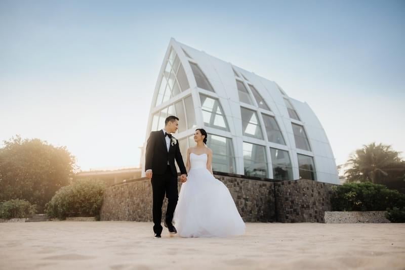  Newlyweds walking hand-in-hand on the sandy beach with the Majestic Chapel at The Ritz-Carlton Bali as a picturesque backdrop 