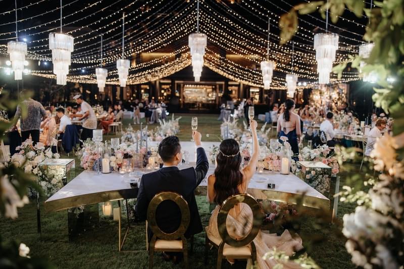  Bride and groom raising a toast under a canopy of twinkling fairy lights and elegant chandeliers during their reception at The Ritz-Carlton Bali's Senses Lawn 