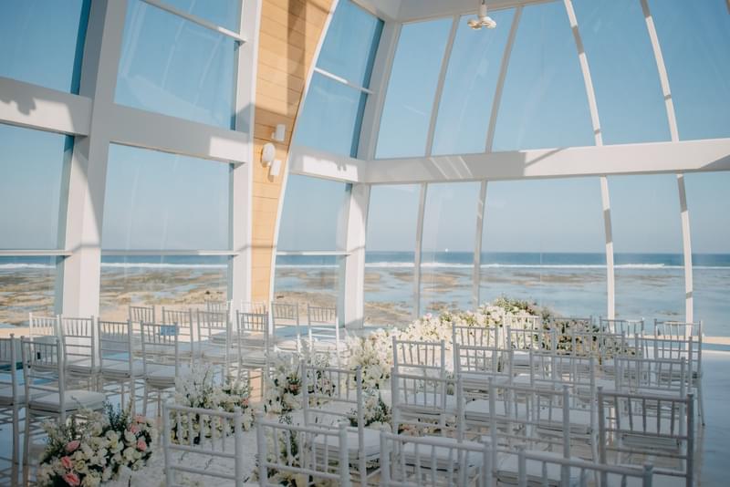  Sunlit interior of the Majestic Chapel at The Ritz-Carlton Bali, featuring elegantly arranged seating and breathtaking ocean views through expansive glass walls 