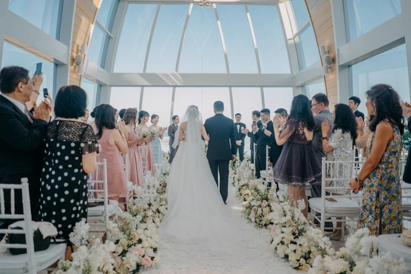  Father and bride walking down the aisle surrounded by loved ones in the Majestic Chapel at The Ritz-Carlton Bali, adorned with elegant floral arrangements and natural light 