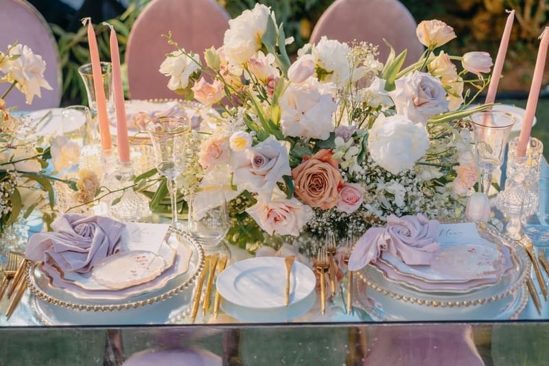  Close-up of an elegant bridal table setting featuring soft pastel floral arrangements, pink taper candles, gold-accented tableware, and luxurious details 
