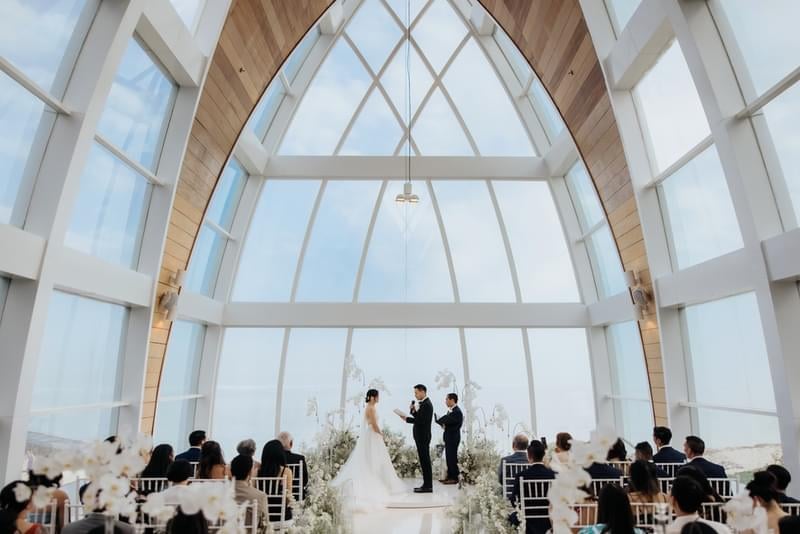  Romantic wedding ceremony inside the Majestic Chapel at The Ritz-Carlton Bali, with floor-to-ceiling windows framing stunning ocean views and natural light 