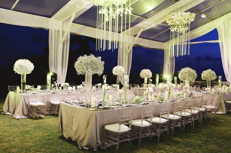  Elegant evening wedding reception setup featuring long banquet tables, lush white floral centerpieces, and cascading chandeliers under a transparent marquee at The Ritz-Carlton Bali 