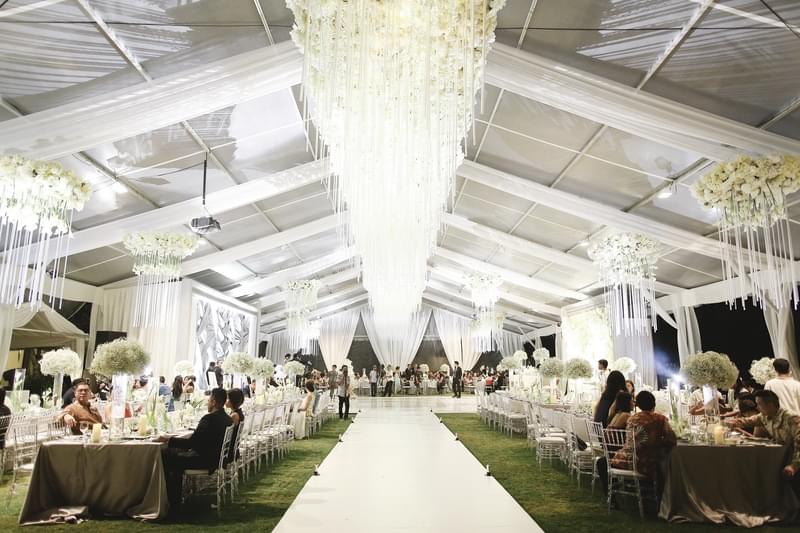  Grand wedding reception under a luxurious marquee at The Ritz-Carlton Bali, featuring a stunning aisle, cascading chandeliers, and elegant white floral decor. 