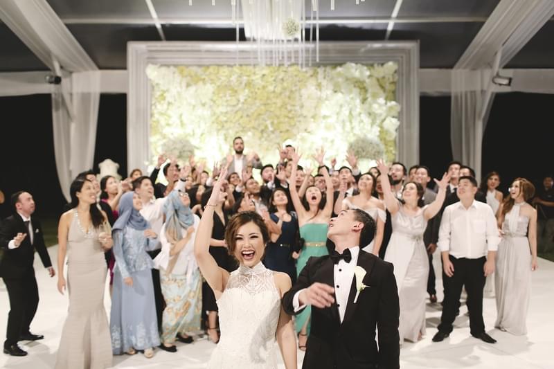  Joyful bride and groom celebrating with their guests under a luxurious marquee at The Ritz-Carlton Bali, surrounded by elegant floral decor and lively energy 