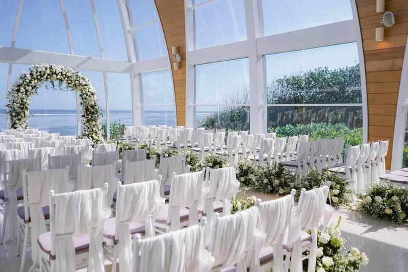  Beautifully arranged wedding seating adorned with white drapes and floral accents inside the Majestic Chapel at The Ritz-Carlton Bali, offering panoramic ocean views 