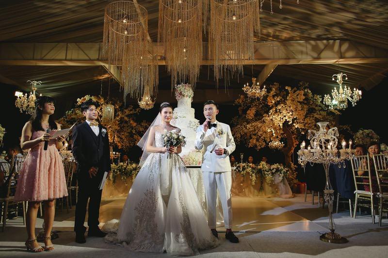  Elegant wedding reception with the bride and groom addressing their guests, surrounded by luxurious chandeliers, a grand wedding cake, and opulent floral decor at The Ritz-Carlton Bali 