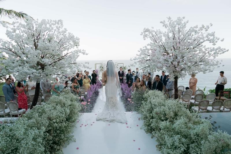  Guests standing amidst cherry blossom trees, watching the bride walk down a floral-lined aisle with a stunning ocean backdrop at The Ungasan 