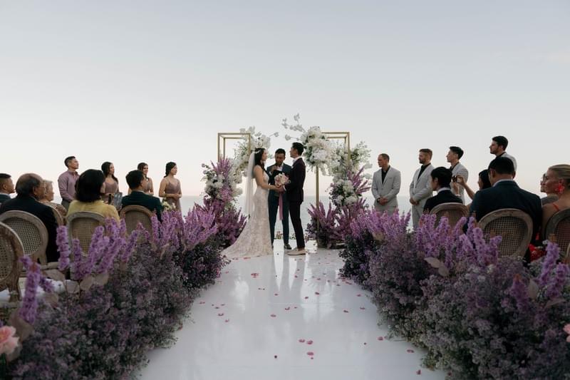  Wedding guests seated in elegant chairs, witnessing a heartfelt ceremony framed by purple florals and a serene ocean view at The Ungasan 