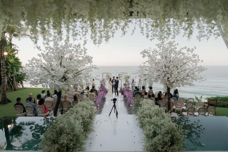  Bride and groom exchange vows at the end of an elegant long aisle surrounded by lush florals and ocean views at The Ungasan 