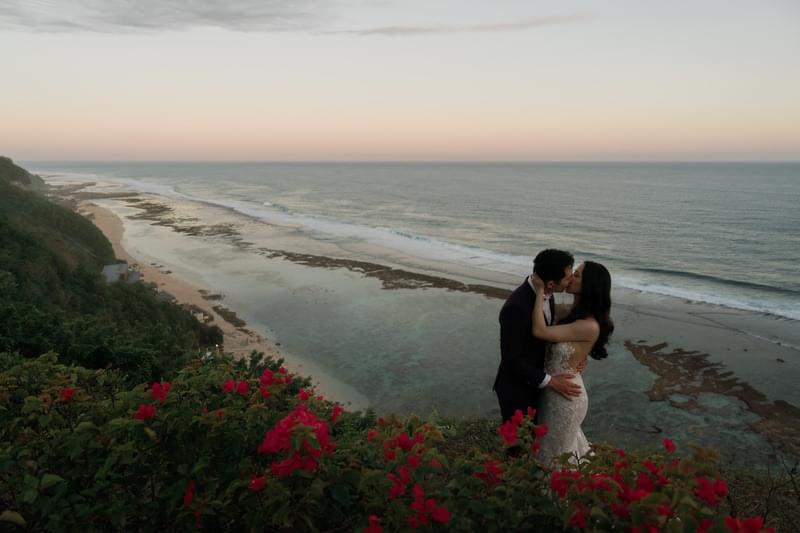  A romantic kiss shared by the bride and groom with stunning clifftop views of the ocean at sunset surrounded by red flowers 
