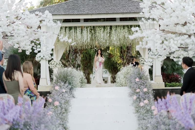  Radiant bride descending a flower-lined aisle under a canopy of white blossoms, making her grand entrance at The Ungasan 