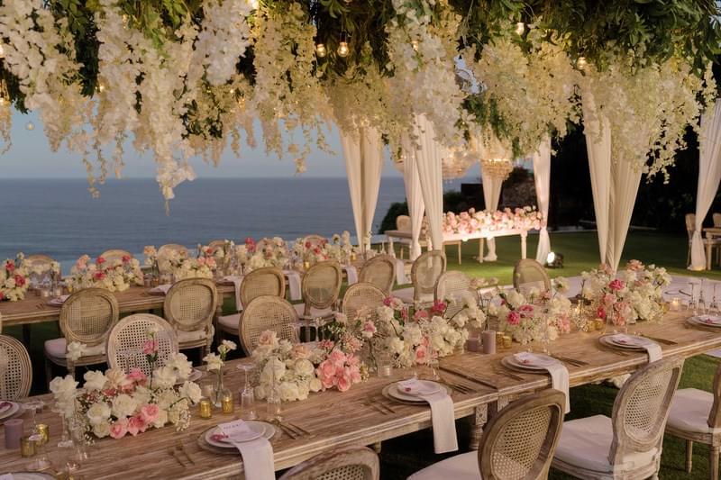  Luxurious outdoor wedding reception setup featuring rustic wooden tables, elegant floral arrangements, and cascading white blooms at The Ungasan 