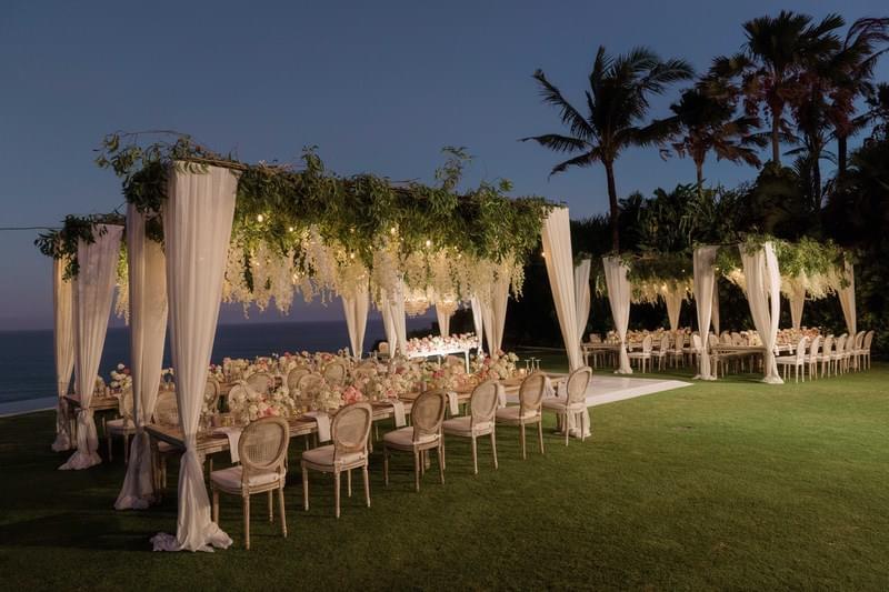  Elegant outdoor wedding reception at The Ungasan, featuring canopy-style floral decor, chic dining tables, and ocean views under the evening sky 