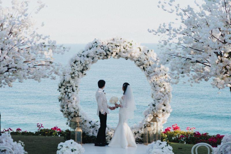 Bride and groom holding hands under a stunning circular floral arch with a breathtaking ocean view at The Ungasan. 