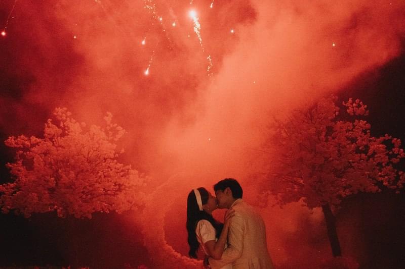  Bride and groom kissing under vibrant fireworks, framed by beautiful cherry blossom trees, at a clifftop wedding reception at The Ungasan 