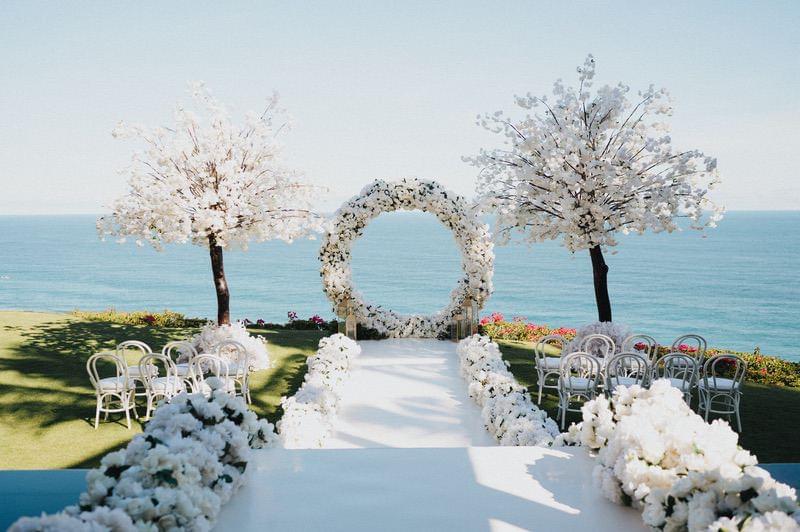  Elegant wedding setup at The Ungasan, featuring white floral arrangements, a circular floral arch, and cherry blossom trees with a breathtaking ocean backdrop 