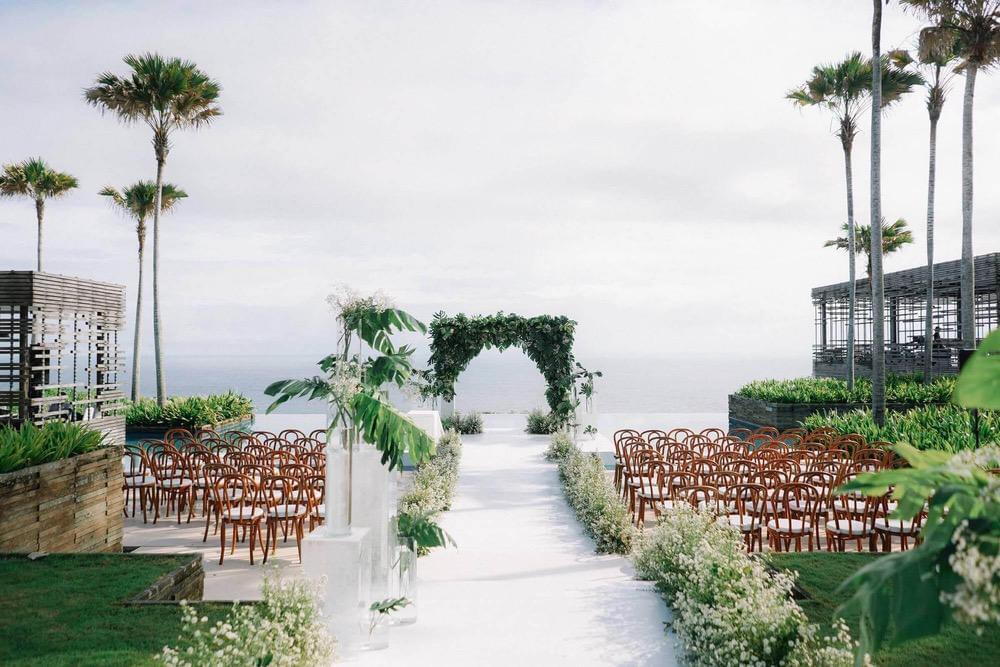 Elegant wedding ceremony setup on Alila Uluwatu's floating stage, featuring a lush green arch, rows of wooden chairs, and expansive ocean views framed by palm trees