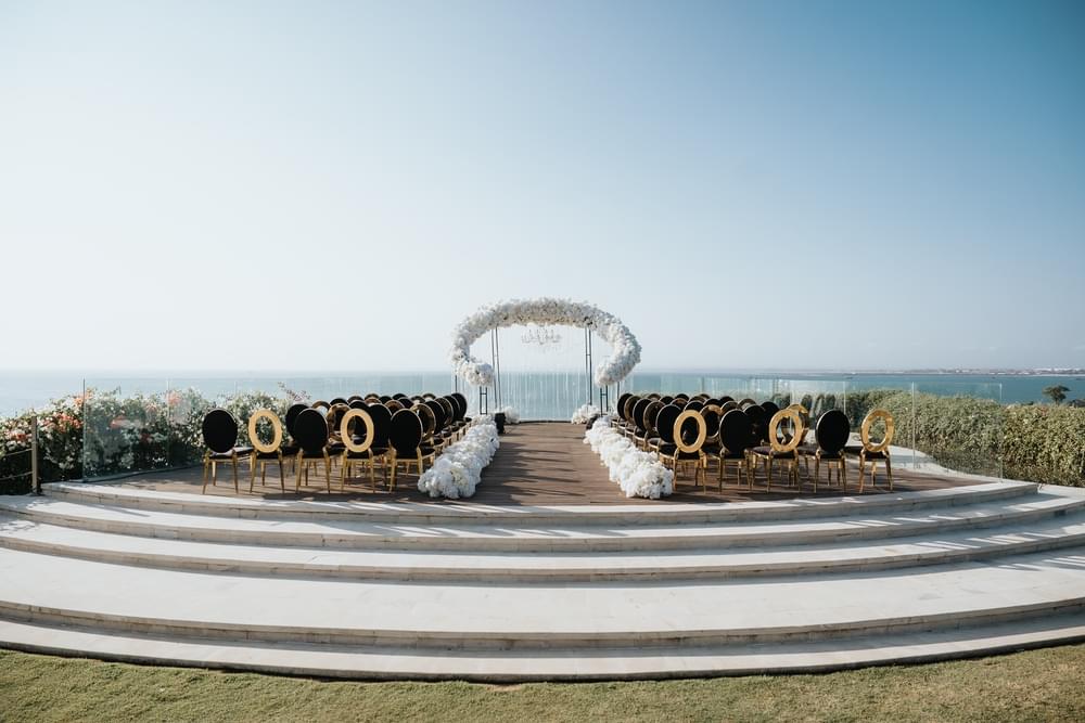 Luxurious wedding ceremony setup on Ayana Villa's cliff-edge platform, featuring black and gold chairs, a flower-lined aisle, and a floral arch with chandelier, overlooking panoramic ocean views.