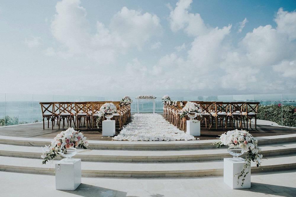 Elegant wedding ceremony setup on Ayana Villa's cliff-edge platform, featuring a flower-lined aisle, rustic wooden chairs, and breathtaking ocean views under a bright sky