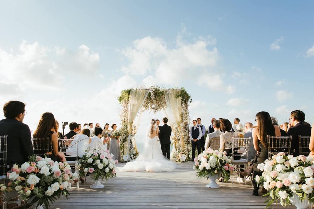 Romantic wedding ceremony at Ayana Villa's cliff-edge platform, featuring a floral-covered canopy, elegant floral arrangements, and guests seated under a bright blue sky with ocean views.