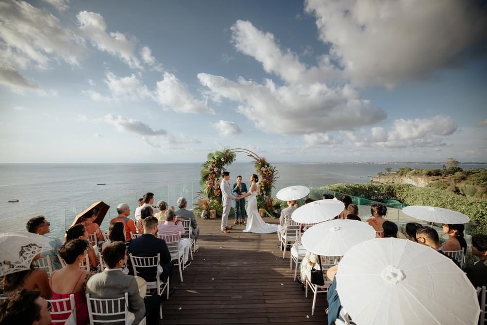 Romantic wedding ceremony on Ayana Villa's cliff-edge platform, featuring white chairs, parasols, and a floral arch, with guests overlooking expansive ocean views under a bright, partly cloudy sky.