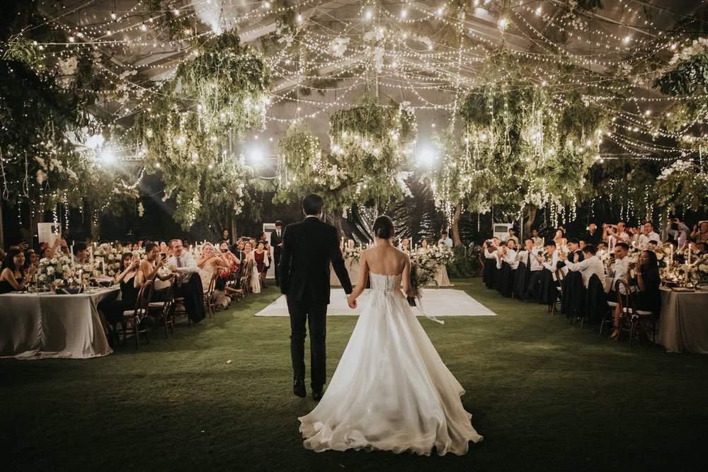 Enchanting garden wedding reception at Ayana Villa, with a bride and groom entering under a canopy of string lights and greenery, surrounded by guests seated at elegantly decorated tables.