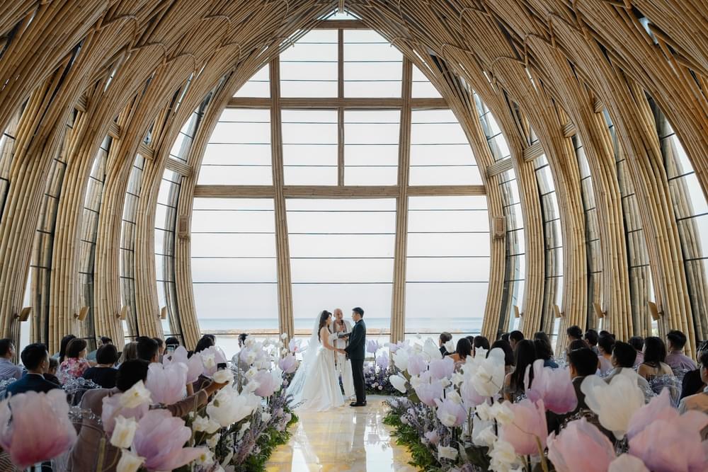 Wedding ceremony at The Apurva Kempinski’s Majestic Chapel, with the couple exchanging vows under towering bamboo arches, surrounded by guests and pastel floral arrangements, overlooking ocean views.