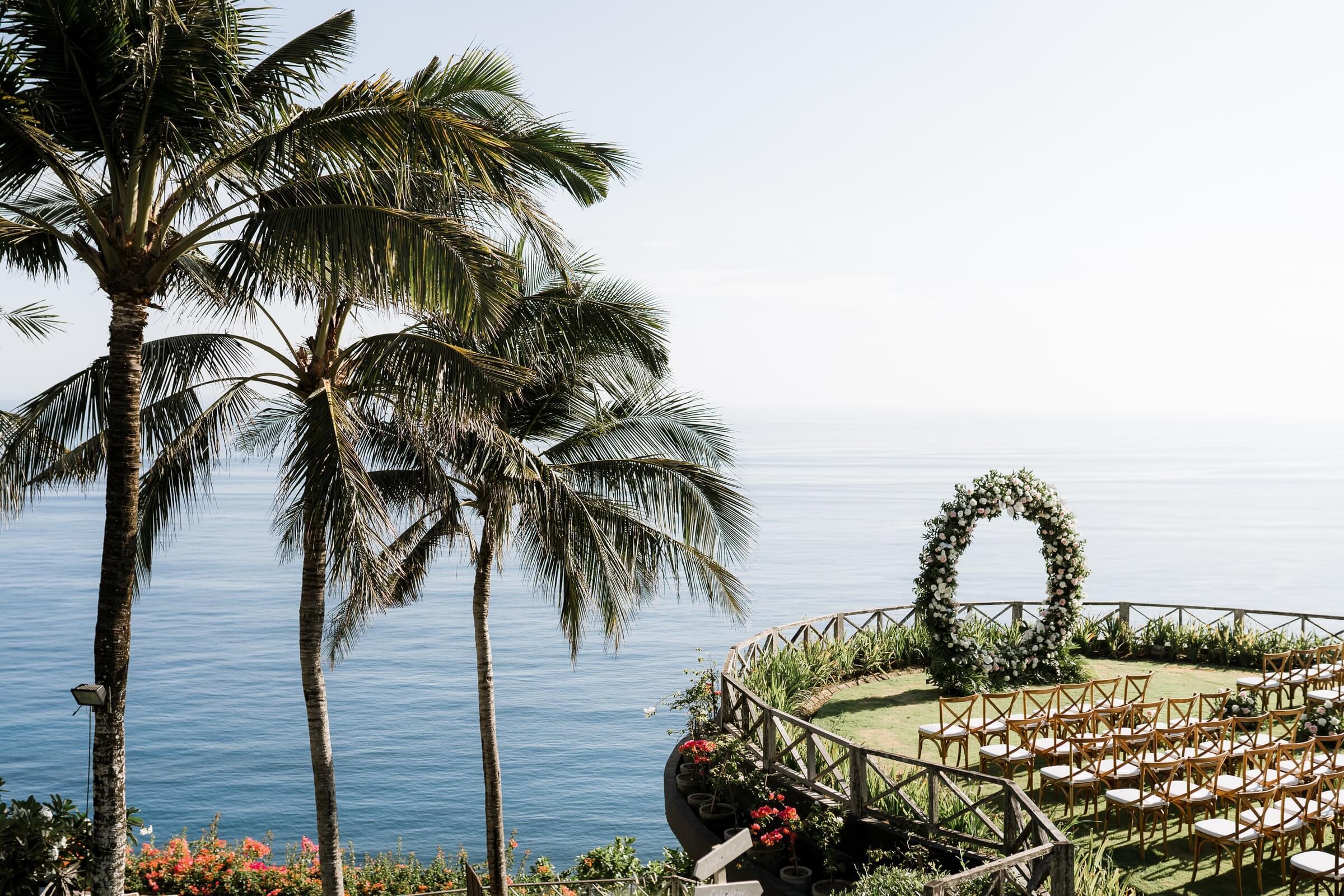 Clifftop wedding ceremony at Khayangan Estate with a circular floral arch, rustic chairs, and stunning ocean views framed by tall palm trees.