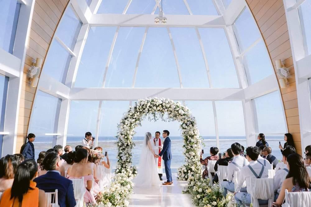 Bride and groom standing under a floral arch in the Majestic Chapel at Ritz Carlton Bali, with guests seated and ocean views visible through large glass windows.