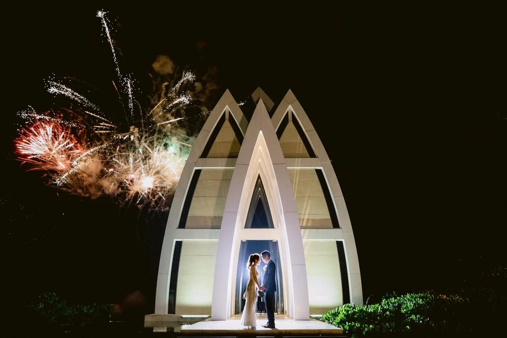 Romantic couple's portrait in front of the Majestic Chapel at The Ritz Carlton Bali, with vibrant fireworks illuminating the night sky.