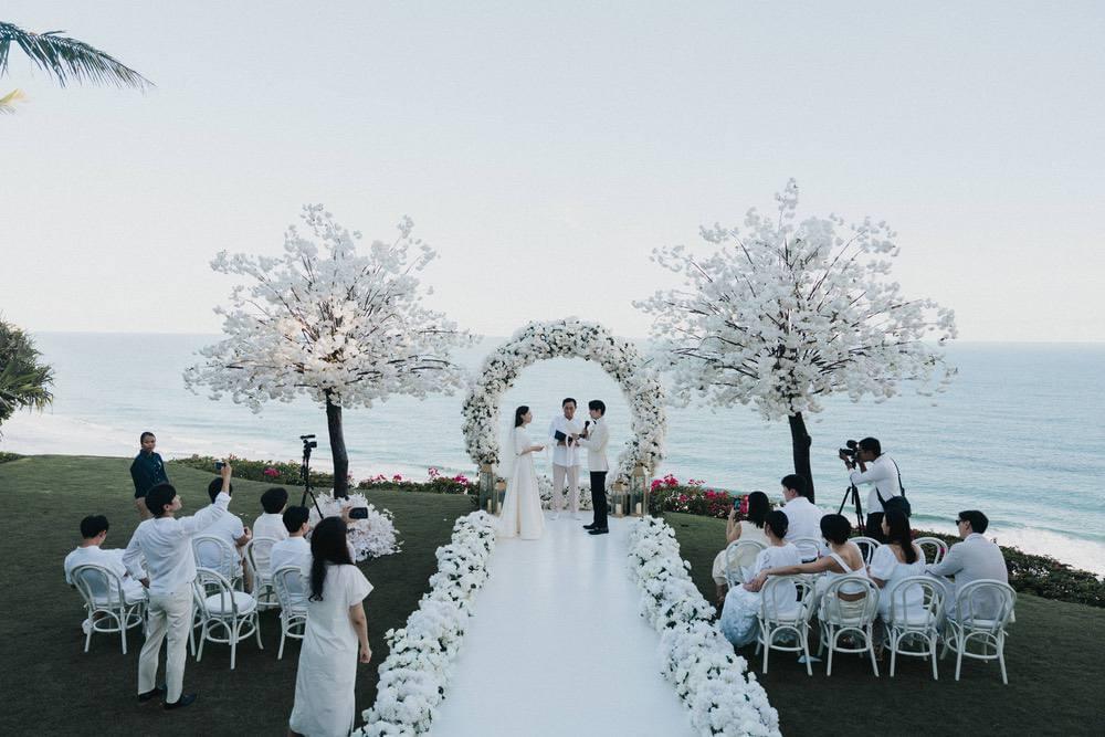 Couple exchanging vows on a white asile surrounded by friedns with ocean backdrop